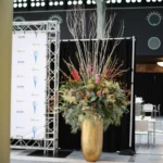 Gouden smalle hoge vaas met witte lange takken en een onderstroom van groene bladeren.