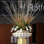 Kunstzinnig gedecoreerde witte vaas op een statief met witte takken en groene bladeren met witte bloemen