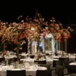 Feestelijke aankleding van ronde tafels met wit tafelkleed, voorzien van diverse hoge bloemstukken op stam.