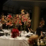 Feestelijke aankleding van ronde tafels met wit tafelkleed, voorzien van diverse hoge bloemstukken op stam.